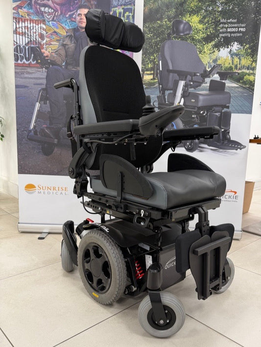 Black electric wheelchair in a showroom setting with promotional materials in the background.